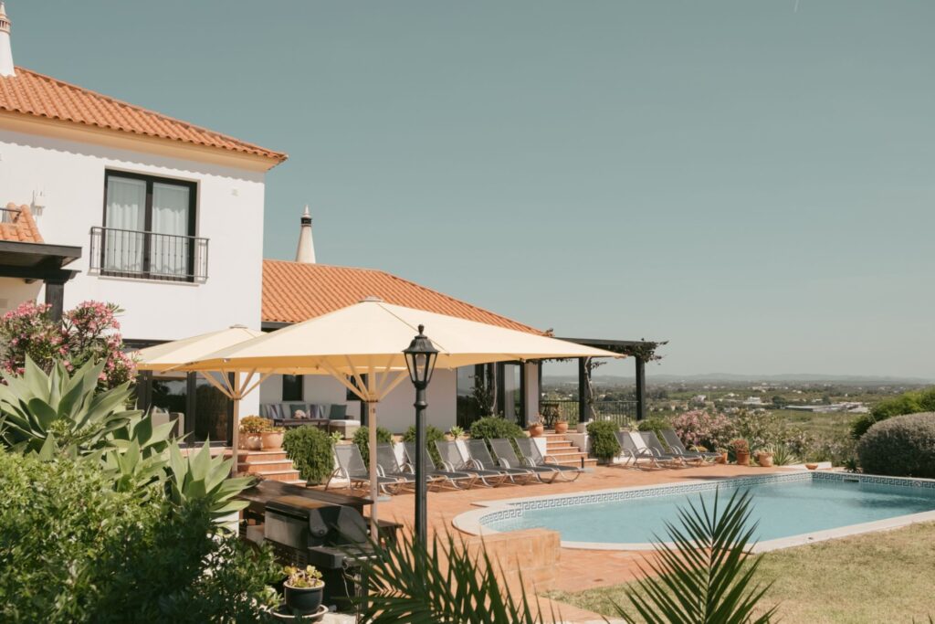 House with swimming pool - POMAR na praia Mediterranean-style white villa at POMAR na praia with terracotta roof, shady umbrellas, sun-loungers and a turquoise swimming pool overlooking the Algarve countryside.