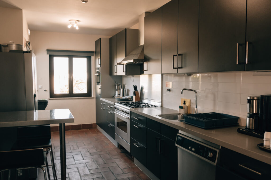 Kitchen - POMAR na praia Modern fully equipped shared kitchen at POMAR na praia coliving, featuring dark cabinets, stainless steel appliances, and natural light from a large window.