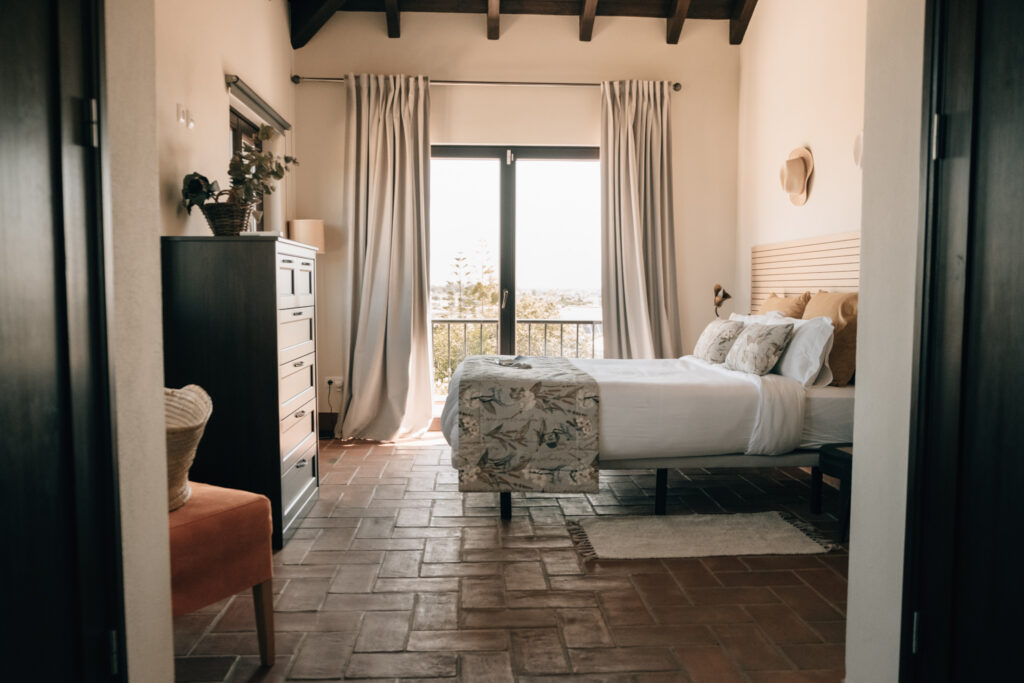 Elegant ground-floor suite at POMAR na praia with large bed, light curtains, terracotta floors, and balcony view.