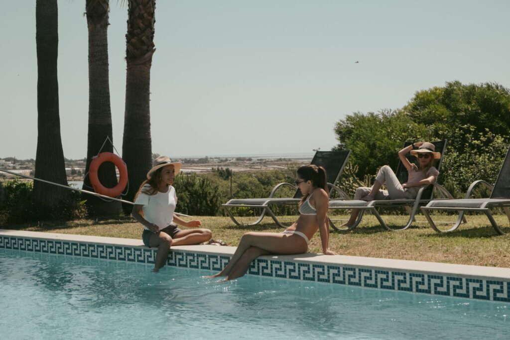 Three women relaxing poolside at POMAR na praia – chatting, dipping feet, and sunbathing under palm trees.
