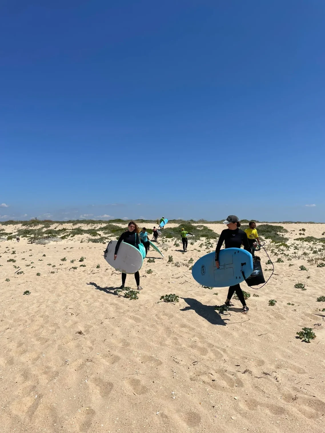 Group of people carrying surfboards across sand dunes toward the ocean