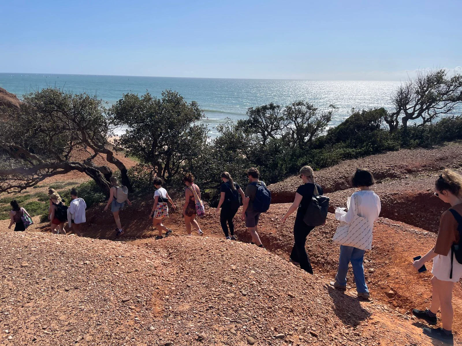 A group walks along a rocky coastal trail with low shrubs and ocean beyond.