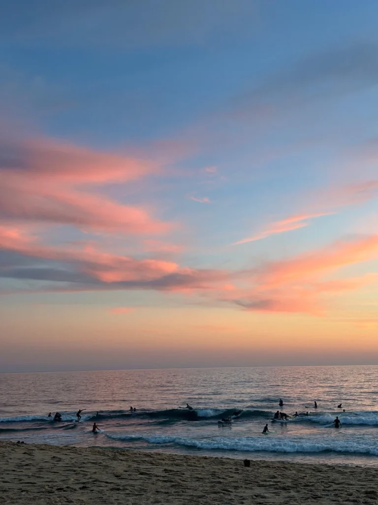 Several people standing in shallow water beneath a pastel sky at a calm ocean beach.