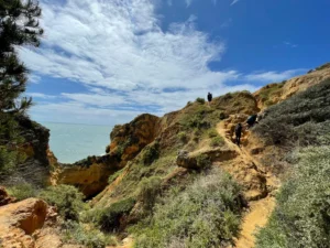 Small hikers follow a rocky coastal hillside path above blue-green water.