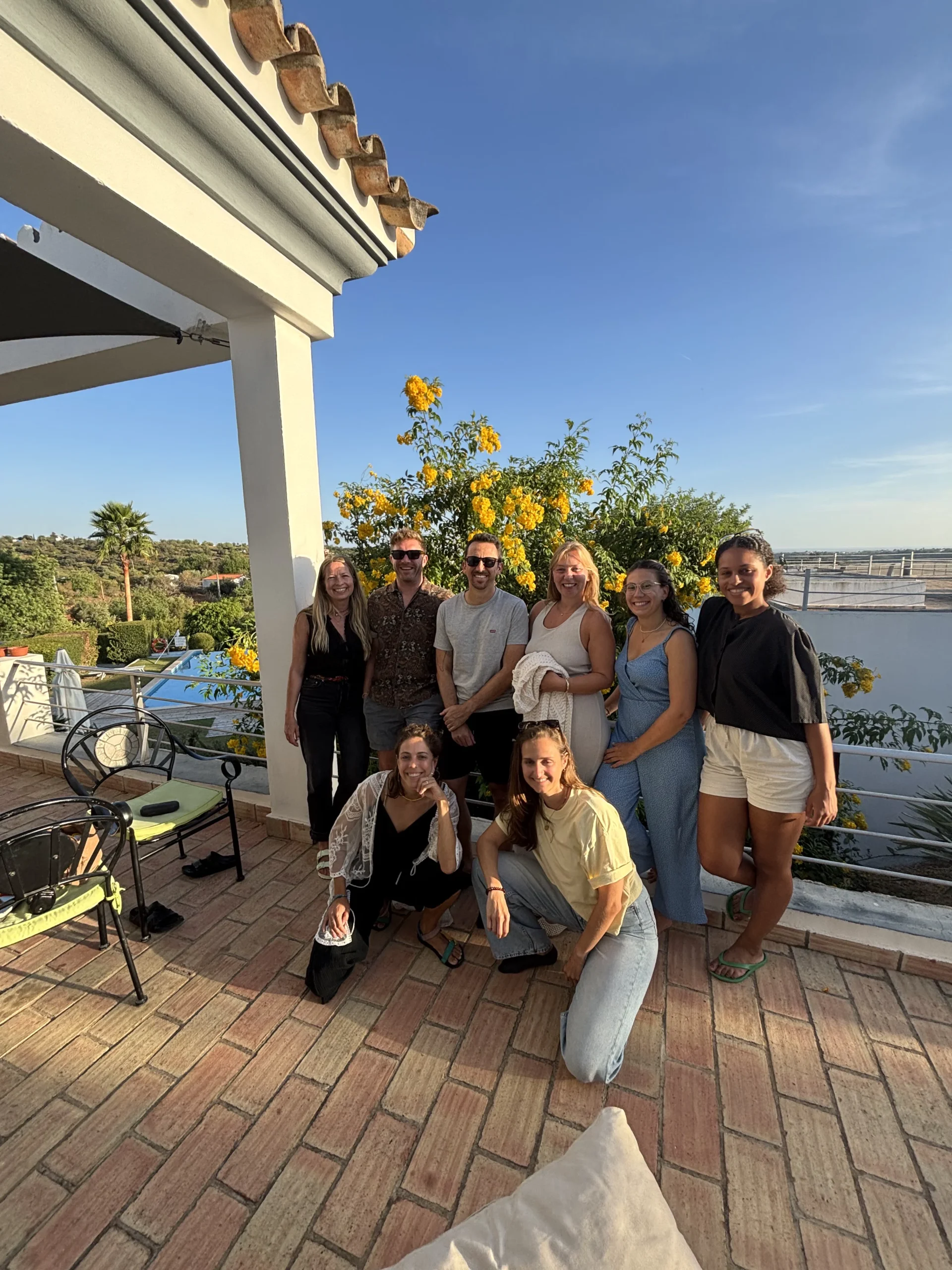 A group gathers on a tiled outdoor terrace beside a white stucco building, with a table, plants, and a broad waterscape.