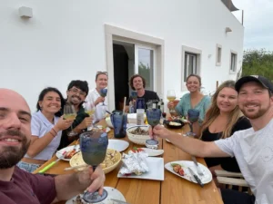 Adults share food and drinks around a wooden outdoor table beside a white building.