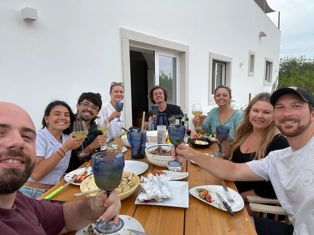 Adults share food and drinks around a wooden outdoor table beside a white building.