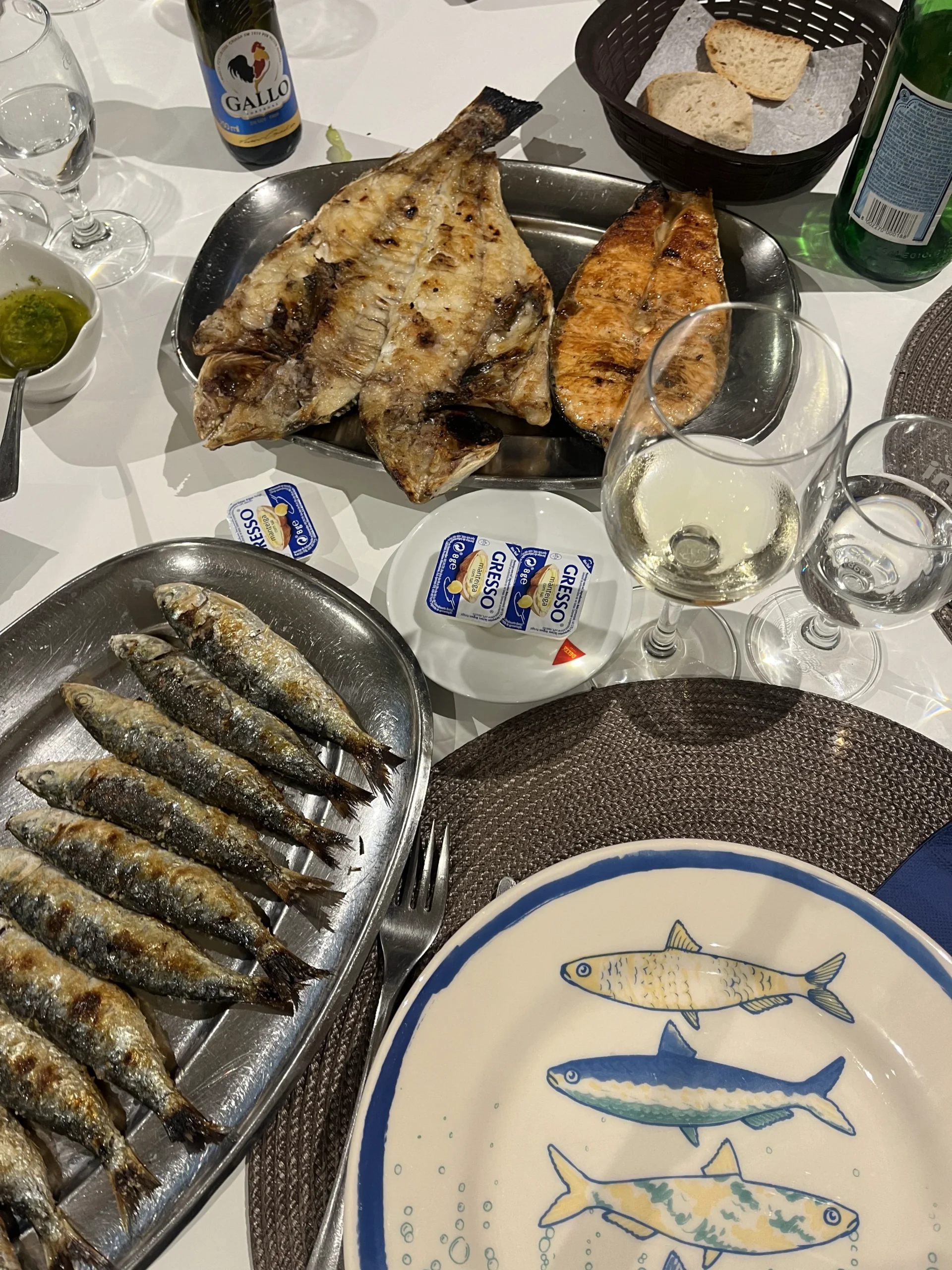 Grilled fish dishes, bread, condiments, and glasses arranged on a dining table.