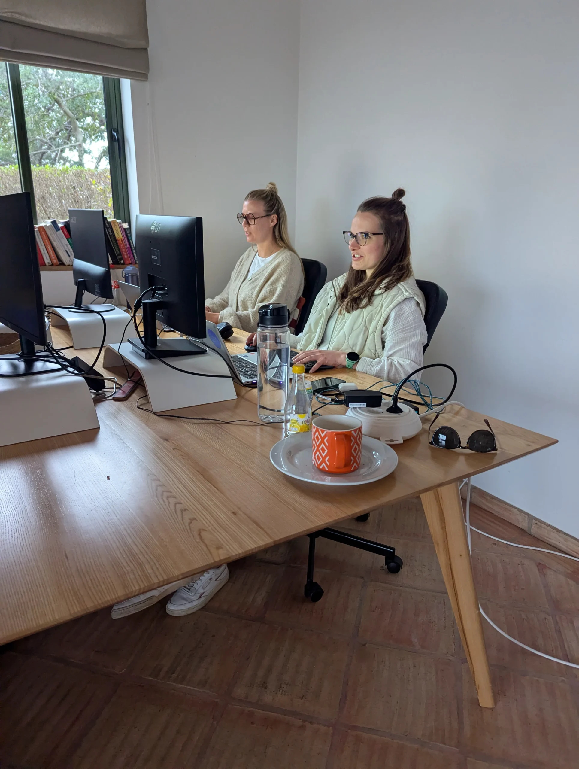 Two people working at computers at a shared wooden desk in a bright office.
