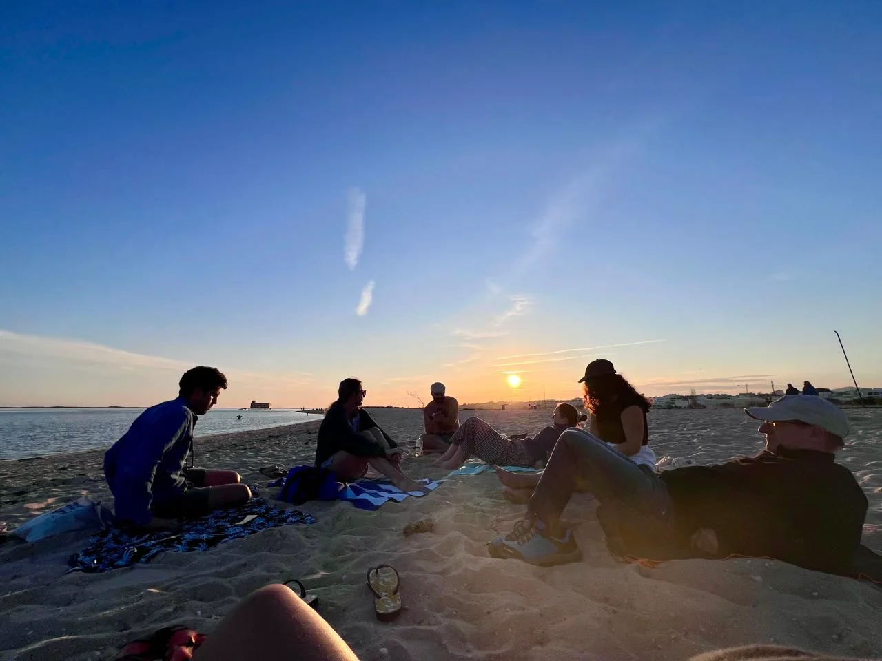 A group sits together on a sandy beach beside the water at sunset.