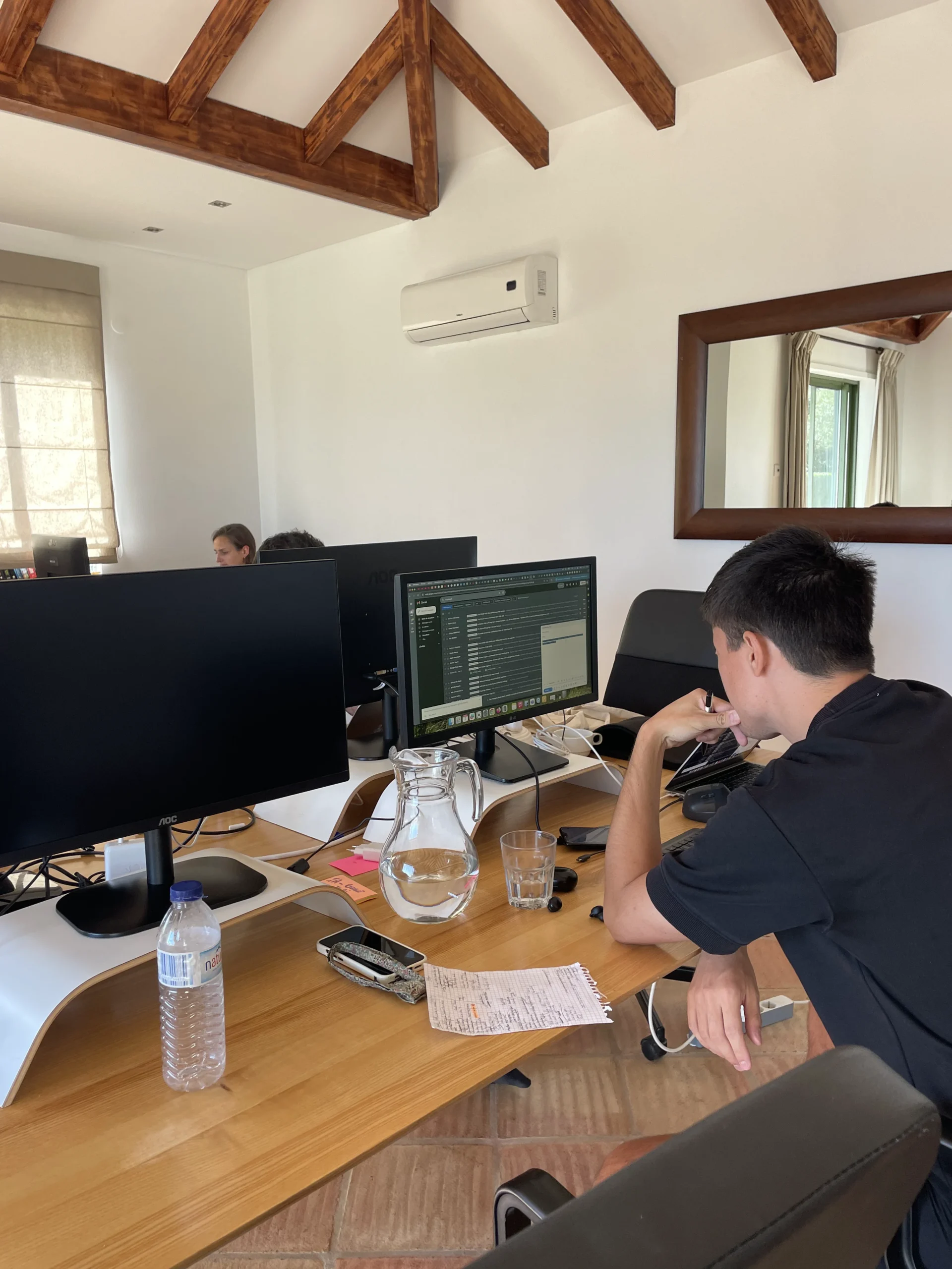 Two people work at a large wooden desk with computer monitors in a bright indoor workspace with exposed wooden ceiling beams.