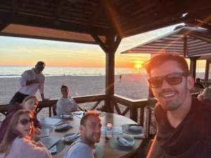 People gathered around an outdoor dining table beneath a seaside pavilion at sunset, with the ocean beyond.