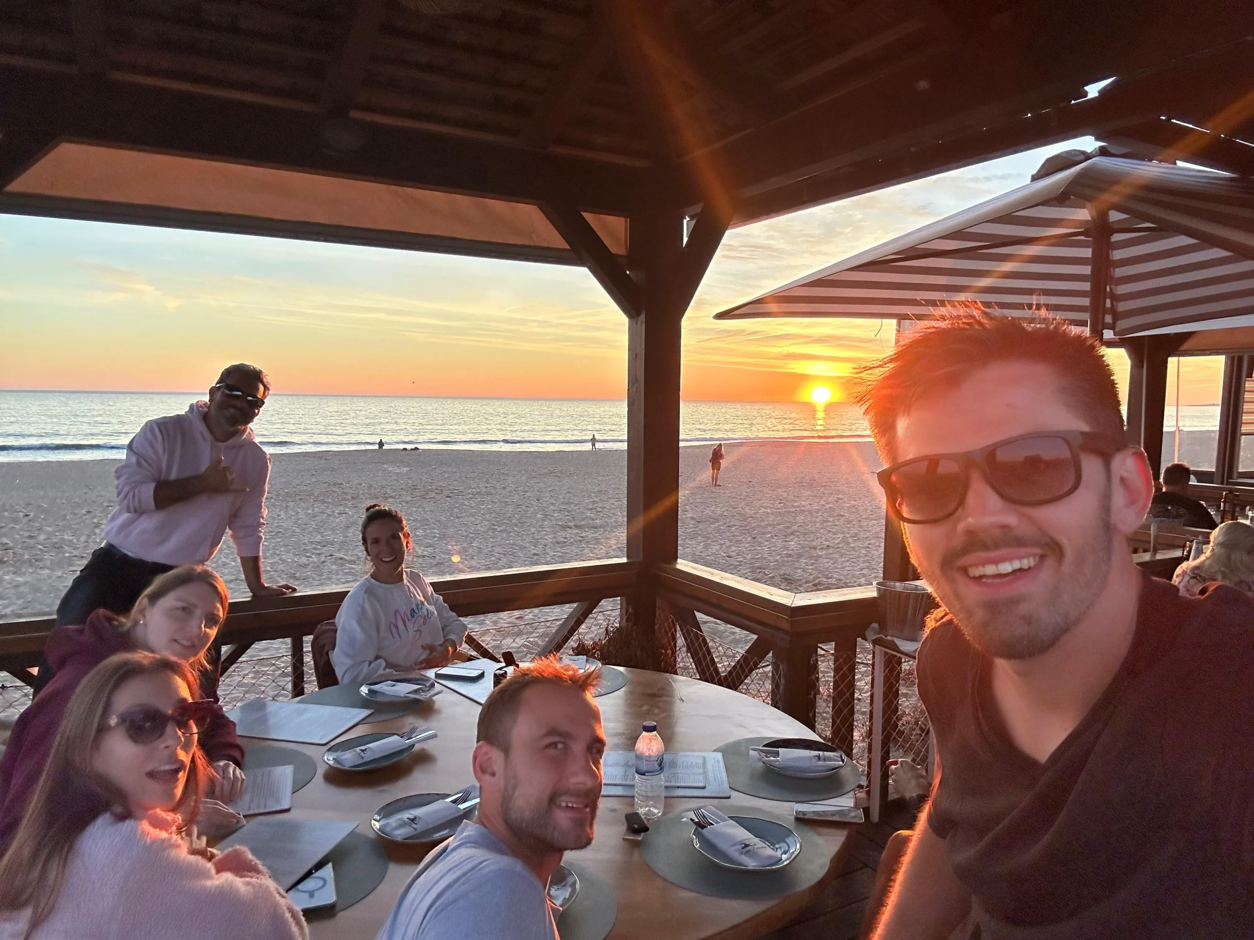 People gathered around an outdoor dining table beneath a seaside pavilion at sunset, with the ocean beyond.