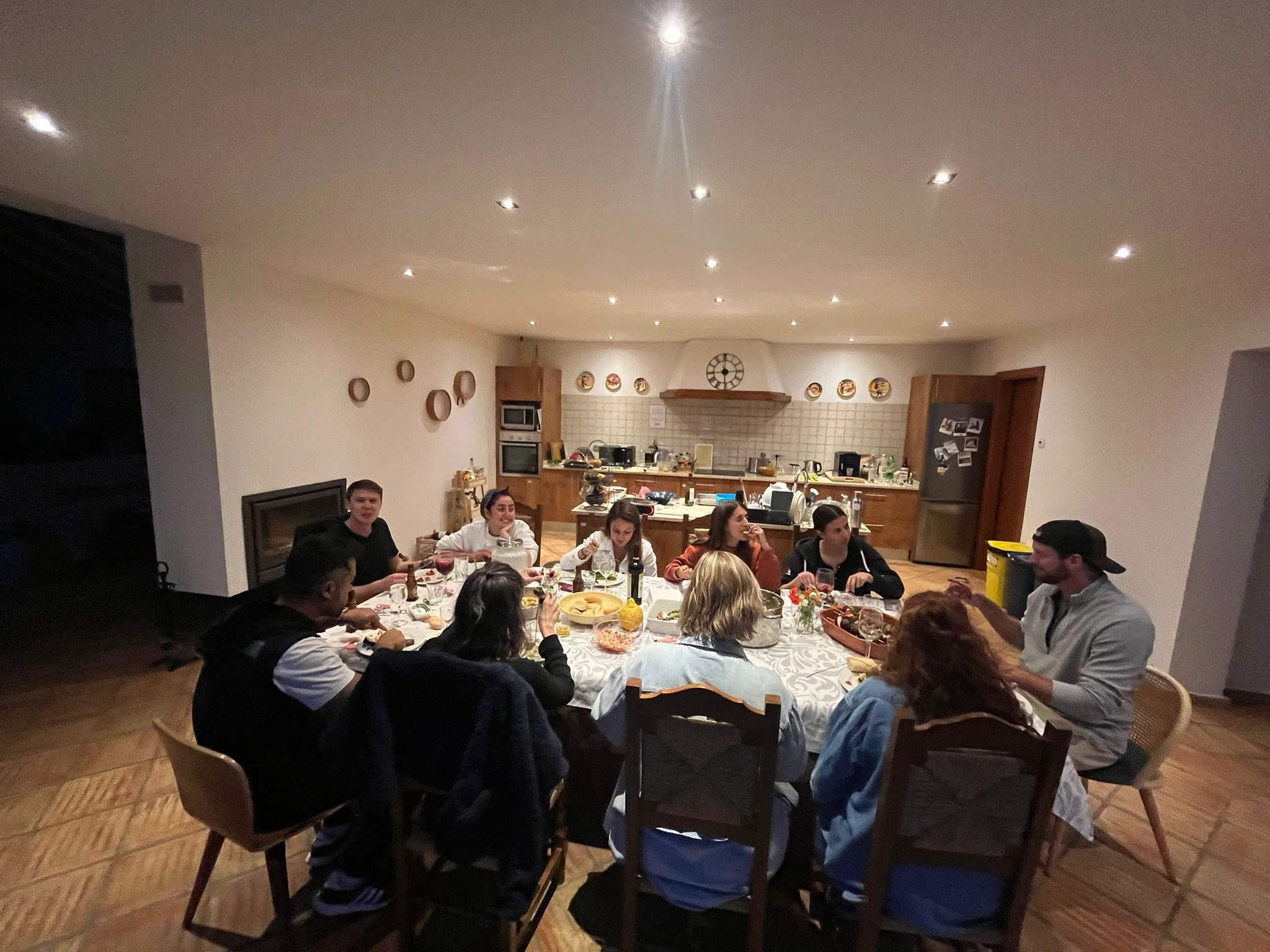 A group shares a meal around a long table in an open-plan kitchen and dining area.