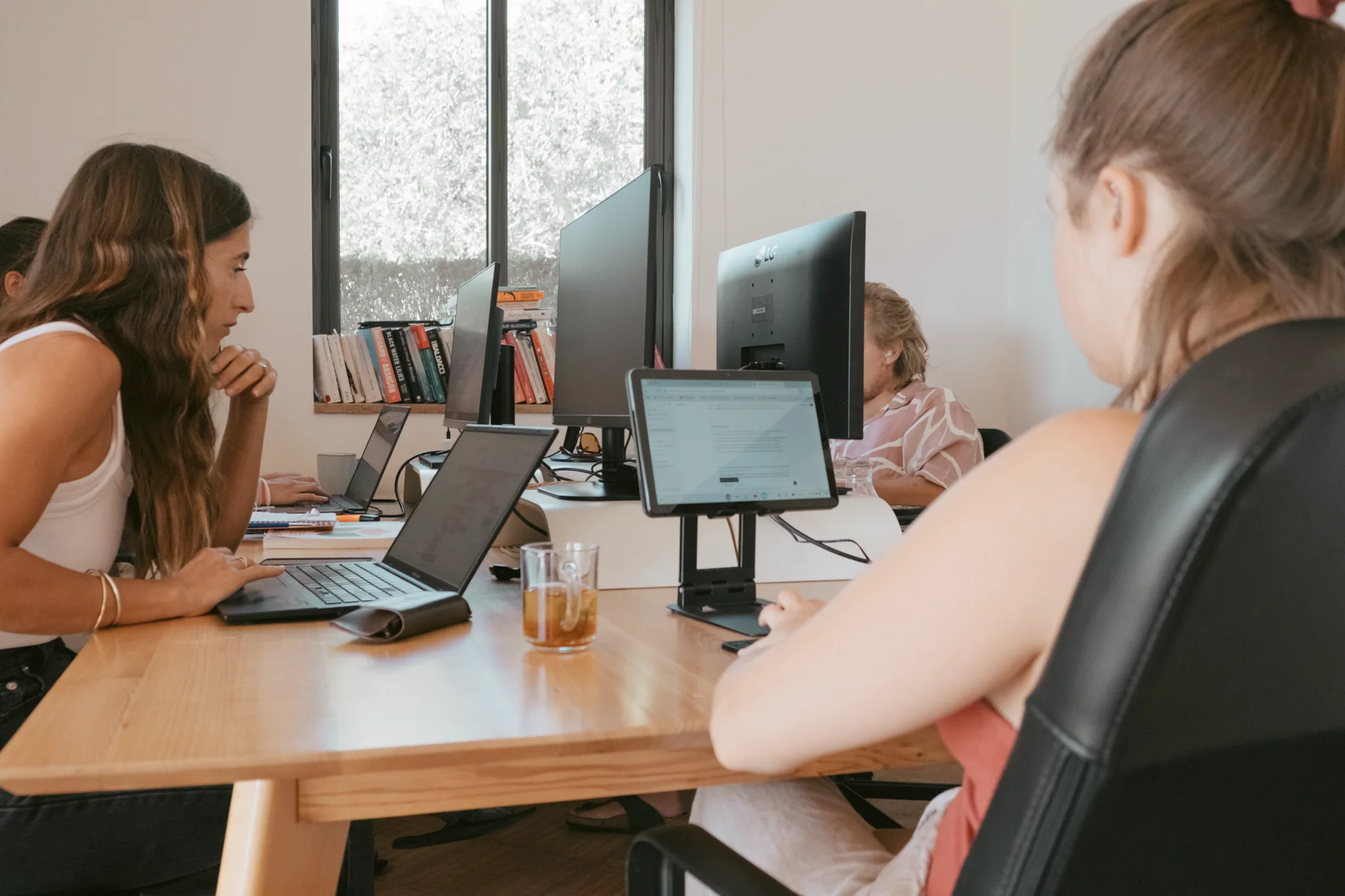 Several people work at computers along a shared desk in a bright office with bookshelves and large windows.